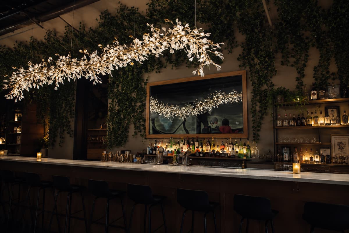 The marble bar at Bar Filo after dark, lit by a hanging brass and crystal fixture with ivy-draped walls and a backlit bottle wall
