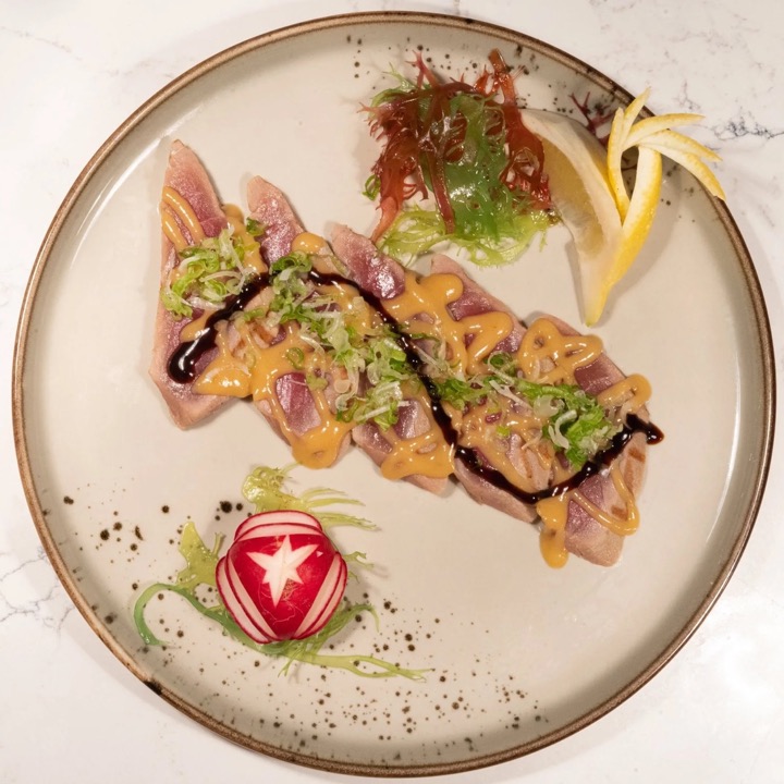 Hamachi carpaccio dressed with yuzu-ponzu and spiced aioli, finished with fried leeks, seaweed and a carved radish, seen from above on a round plate